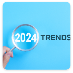 The image features a hand holding a magnifying glass over the text '2024 TRENDS' against a light blue background. The numbers '2024' are prominently displayed in white, while the word 'TRENDS' is in bold black, creating a striking contrast. The use of the magnifying glass suggests a focus on examining or exploring upcoming trends for the year 2024, conveying a sense of curiosity and analysis. The overall design is clean and modern, emphasizing clarity and attention to detail.