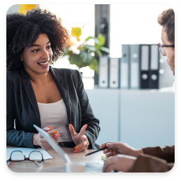 The image shows a professional setting where two individuals are engaged in a conversation. A woman with curly hair is animatedly speaking, wearing a blazer and a white top, suggesting a business context. She appears confident and expressive, using hand gestures to emphasize her points. The man across from her, wearing glasses, is listening attentively, indicating a collaborative discussion. The background features office elements, such as file organizers, contributing to the professional atmosphere. The lighting is bright, enhancing the focus on their interaction.