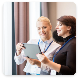 The image shows two women engaged in a conversation while looking at a tablet. The woman on the left has blonde hair and is wearing a light blue blouse, smiling as she interacts with the device. The woman on the right has dark hair and is dressed in a black top, also smiling and appearing interested in what they are viewing together. They are in a well-lit environment, likely at a conference or professional event, with large windows in the background. Both women are wearing lanyards, indicating they are participants in the event. The overall atmosphere is collaborative and positive, suggesting they are sharing ideas or information.