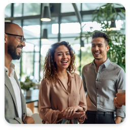 The image features a lively group of three individuals engaged in a friendly conversation. They are in a bright, modern setting with large windows and greenery in the background, suggesting a relaxed and inviting atmosphere.